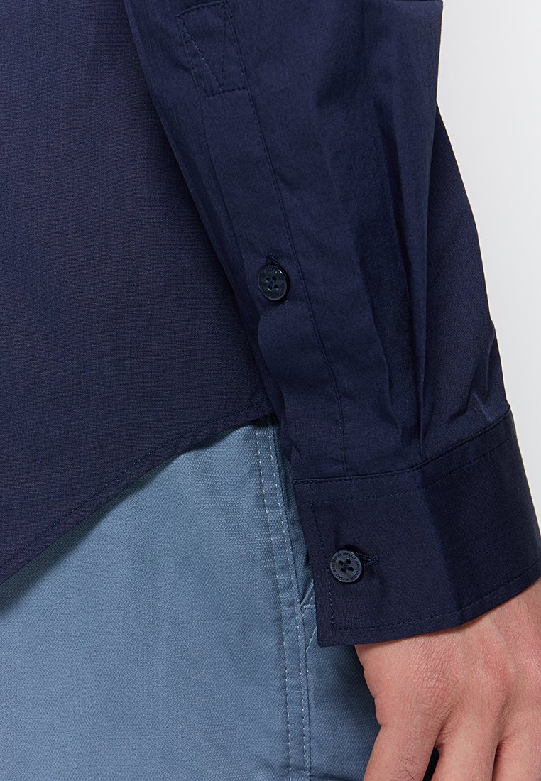 Chemise bleue marine à boutons, avec une finition texturée, présentant des poignets arrondis et deux boutons noirs. Le tissu semble lisse et structuré.