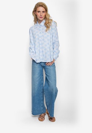 Jasnoniebieska koszula na guziki w okrągłe wzory, zestawiona z szerokimi, dżinsowymi spodniami o luźnym kroju oraz beżowymi sandałami na cienkich paskach.
