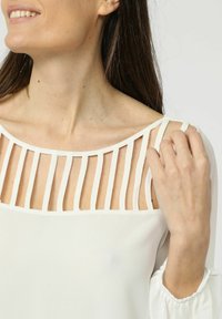 Blusa blanca con escote redondeado y cortes verticales, y delicadas correas. Las mangas son ligeramente acampanadas con puños fruncidos.