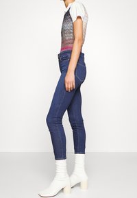 Camiseta gráfica con un diseño degradado en tonos de verde, rosa y blanco, combinada con unos jeans ajustados de color azul oscuro y botines de tacón blanco.