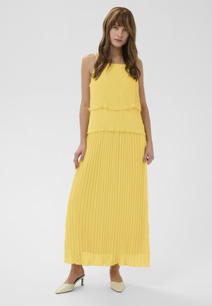 Young woman wearing a sleeveless, yellow pleated maxi dress and beige low-heeled shoes, standing against a plain light background.