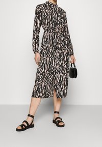 Svart och rosa mönstrad klänning med hög hals, långa ärmar, knytmidja och slits; som matchas med svarta sandaler med remmar och en liten handväska.