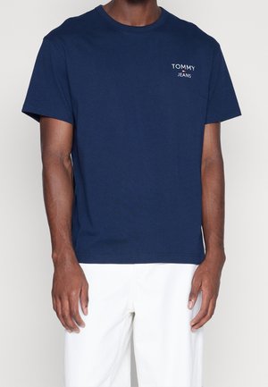 Mężczyzna w granatowym t-shircie Tommy Jeans i białych spodniach, stojący na tle jasnego, jednolitego tła.