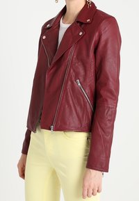 Donna che indossa una giacca da biker attillata in pelle bordeaux con zip argento e pantaloni giallo pallido, in piedi davanti a uno sfondo bianco.