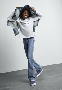 Jeune personne souriante mettant une veste en denim, portant un t-shirt blanc Levi's, un jean bleu et des baskets bleues avec des lacets roses.