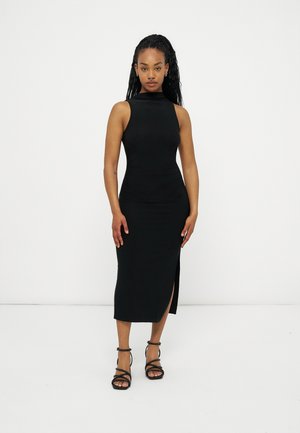 Žena s dlouhými zapletenými vlasy, oblečená v černých midi šatech bez rukávů s bočním rozparkem a černých páskových sandálech na vysokém podpatku, stojící.