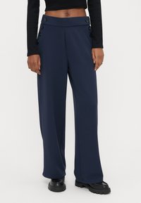 Person iført marineblå vidbukser med høyt liv og knappedetaljer på siden, og svarte kraftige sko, stående mot en ensfarget bakgrunn.