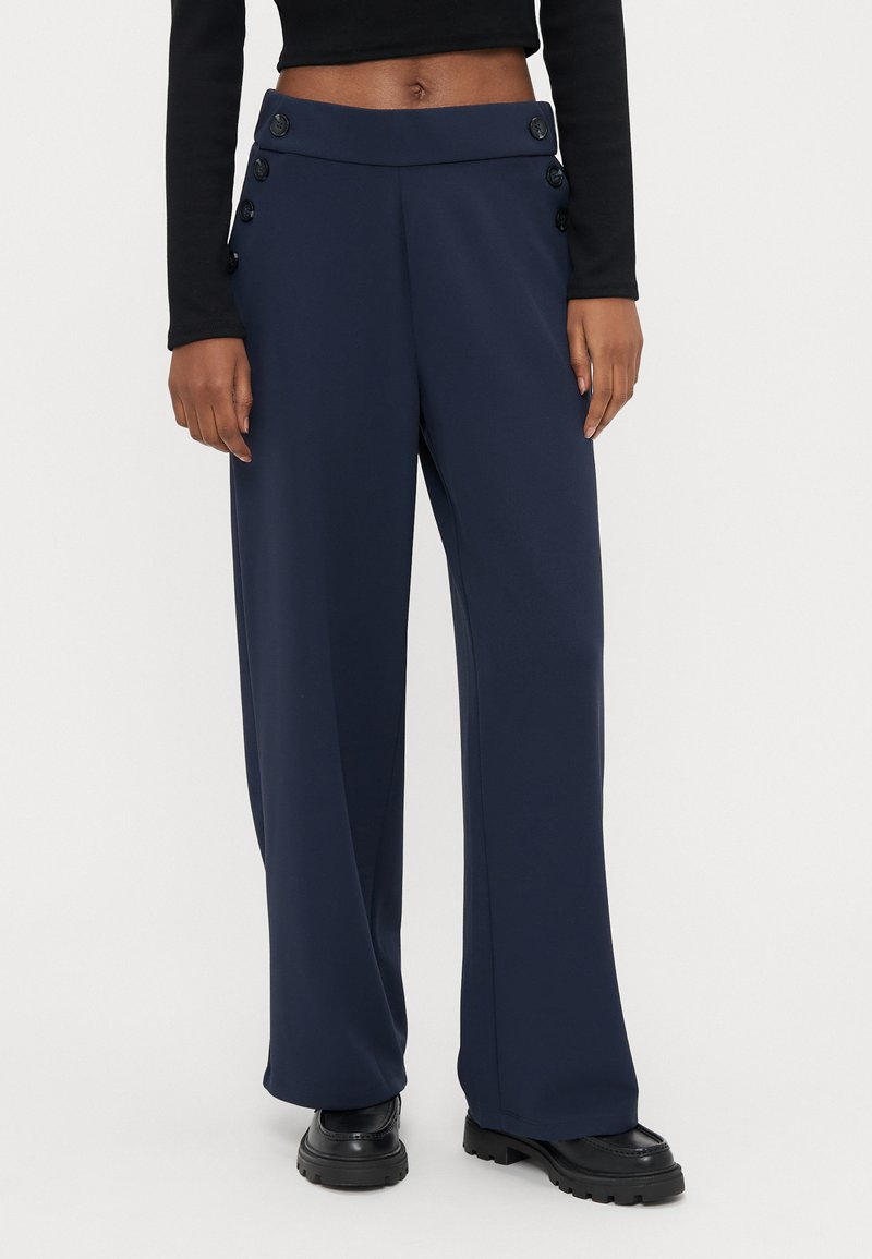 Person iført marineblå vidbukser med høyt liv og knappedetaljer på siden, og svarte kraftige sko, stående mot en ensfarget bakgrunn.