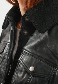 Veste en cuir noir avec col en peau lainée, boutons-pression métalliques, fermeture éclair et poches à rabat, portée sur une chemise blanche.