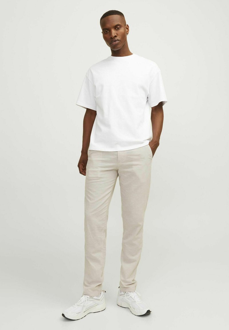 Weißes kurzärmeliges T-Shirt, beigefarbene maßgeschneiderte Hose und weiße Sportschuhe. Der Stoff wirkt weich mit einem lockeren Schnitt und subtilen Nähdetails.