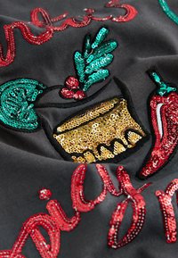 Zwarte stof met kleurrijke paillettenborduursels van limoen, chili en een gouden beker met rode accenten, die een levendig decoratief patroon vormen.