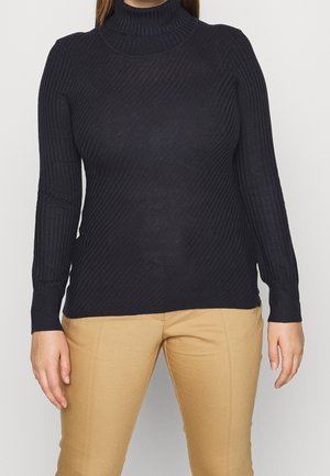 Femme portant un chandail à col roulé noir à côtes et un pantalon beige, se tenant debout devant un fond clair uni avec les bras détendus le long du corps.