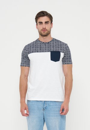 Homme portant un T-shirt blanc avec un motif géométrique bleu marine sur la partie supérieure de la poitrine, les manches et la poche, associé à un jean bleu clair, debout devant un fond blanc.