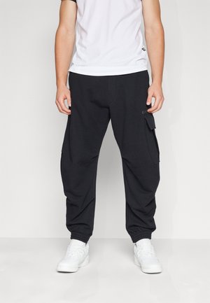 Personne portant un pantalon jogger cargo noir avec poches latérales et poignets élastiques, des baskets blanches et un t-shirt blanc uni, debout contre un mur blanc.