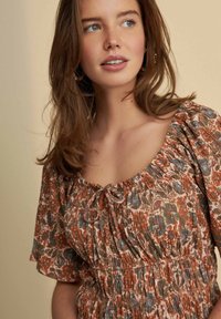 Floral-patterned top with elasticized neckline and waist, featuring short puff sleeves and a bow tie detail at the front. Warm color palette.