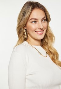Cercei din aur și o brățară din aur răsucită completează un top alb cu dungi fine, evidențiind o textură netedă și un design elegant.