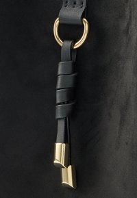 Mustast seemisnahast käekoti detail, millele on kinnitatud keeratud musta nahast rihm kuldse rõnga külge, rihma otstes kuldsed aktsendid.