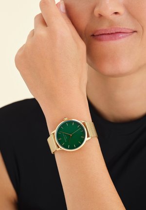Montre verte avec bracelet en maille dorée ; cadran rond, design minimaliste ; possède des index en or et une couronne à la position 2 heures.