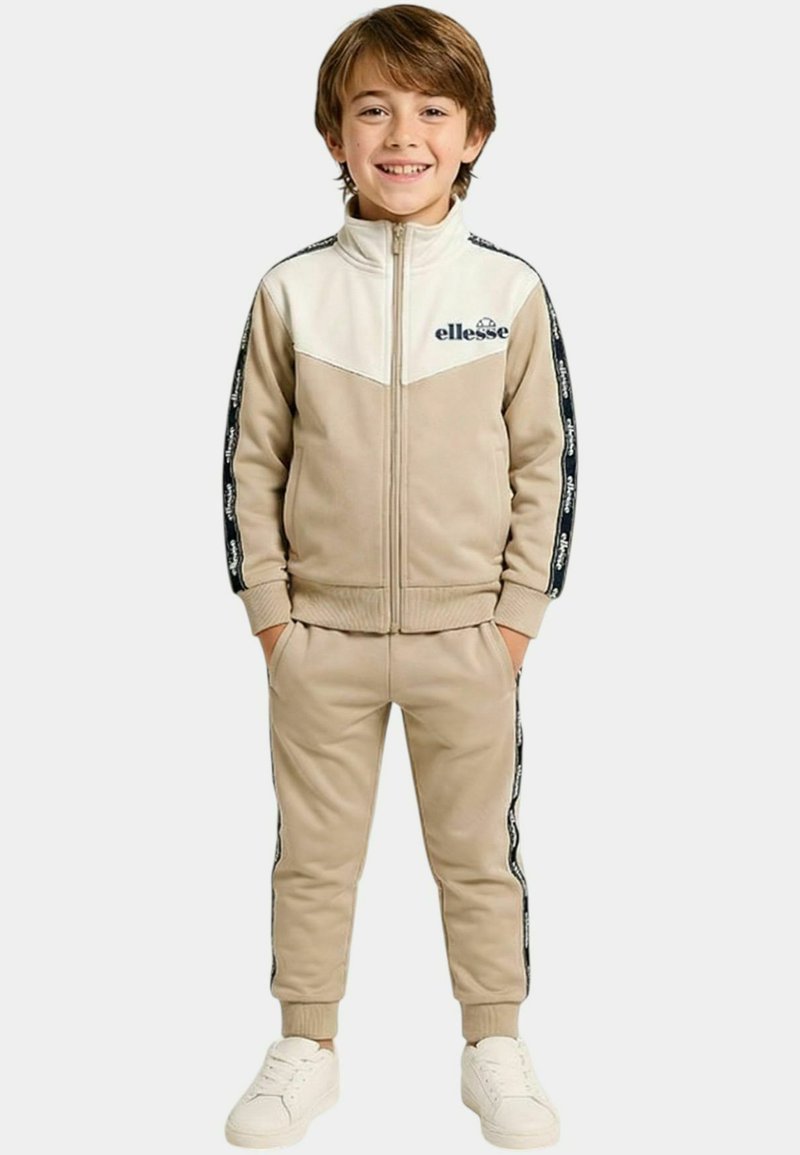 Jeune garçon souriant, portant un survêtement beige et blanc avec des logos à bandes noires sur les manches et les pantalons, baskets blanches, mains dans les poches.