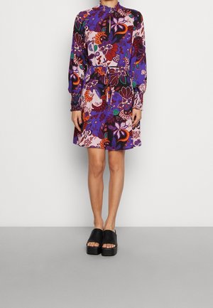 Langes lila Blumenkleid mit langen Ärmeln, hohem Kragen, Taillenkordel und gerafften Manschetten; kombiniert mit schwarzen Plateausandalen.