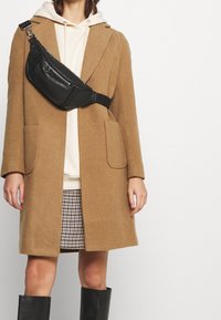 Abrigo de lana color camel con grandes bolsillos, bolso cruzado de cuero negro, sudadera ligera, falda de patrón de pata de gallo y botas negras hasta la rodilla.