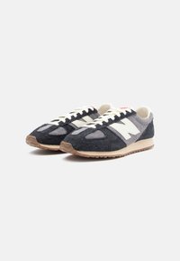 Suedesneakers med en overdel som hovedsakelig er svart, grå tekstilaccent, hvit logo og lisser, samt en tan gummisåle. Teksturt finish.