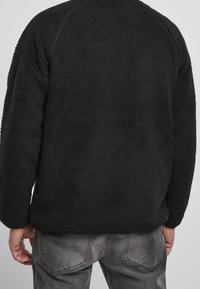 Schwarzer Fleece-Pullover mit einer strukturierten, weichen Oberfläche. Mit abgerundetem Saum und Raglanärmeln, kombiniert mit grauen Jeans.