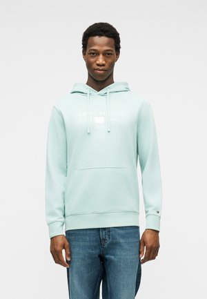 Fiatal férfi világoskék Tommy Hilfiger kapucnis pulóverben és kék farmernadrágban, előre nézve, kezei oldalt lazán leengedve.