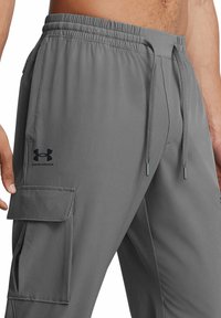 Graue Cargo-Shorts mit elastischem Bund und Kordelzug, ausgestattet mit einer Seitentasche und einem schwarzen Under Armour-Logo am linken Oberschenkel.