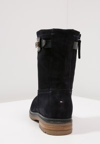 Mörkblå mocka ankelsko med ett svart rem och en metallspänne som accenter. Har en texturerad sula och en liten logotyp nära hälen.