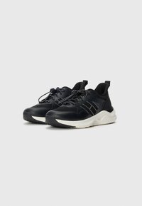 Chaussures de sport noires avec une tige texturée, semelle blanche contrastante et lacets noirs intégrés. Elles disposent d'une languette à l'arrière.