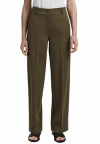 Femme portant un pantalon large taille haute vert olive avec plis devant, bouton marron, passants de ceinture et sandales noires à bout ouvert.