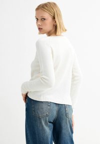 Suéter blanco de punto con cuello redondo, diseño ajustado y dobladillo acanalado. Combinado con jeans de mezclilla azules sueltos, mostrando un look casual.