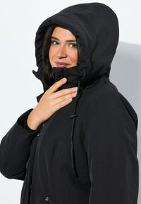 Femme portant une veste à capuche noire, tenant le col près de son menton, avec un léger sourire sur un fond blanc uni.