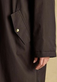 Tuvojoties tumši brūnas mēteļa piedurknes tuvplānam, blakus flapa kabatai, ko tur ar zeltītu pogu.