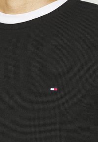 Schwarzes, strukturiertes Strick-T-Shirt mit einem gerippten Rundhalsausschnitt, das ein kleines Logo in Rot, Weiß und Blau auf der linken Brustseite zeigt.