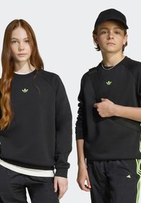 Sweatshirts pretos com mangas raglan, com um pequeno logo verde néon. O garoto usa um boné e segura uma bolsa de ombro, ambos em preto.