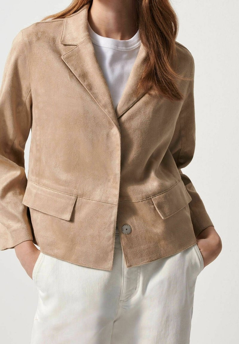 Femme portant un blazer en daim beige sur une chemise blanche, les mains dans les poches d’un pantalon blanc taille haute sur un fond uni.