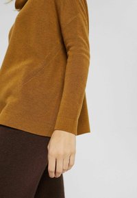 Pull en maille de couleur moutarde avec une texture côtelée et une coupe décontractée, doté d'un col montant et d'un détail de couture diagonale.