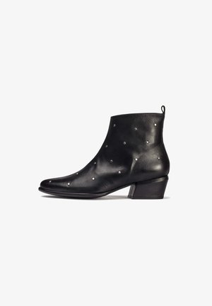 Botín de cuero negro hasta el tobillo con punta afilada, detalles de tachuelas plateadas y un tacón bajo y cuadrado. Textura suave y diseño minimalista.