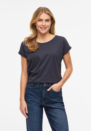 Usmievajúca sa žena s blond vlasmi, oblečená v jednofarebnom tmavosivom tričku s krátkym rukávom a modrých džínsoch, stojaca s jednou rukou v vrecku.