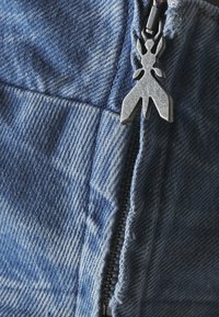 Tejido de denim en azul claro con una textura desgastada. Presenta un tirador de cremallera de metal en forma de insecto y detalles de costura visibles.