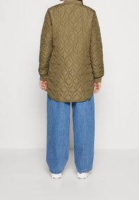 Person i en olivengrøn, kviltet jakke med diamantmønstre, blå bredbenede jeans og hvide sneakers, der står foran en ensfarvet baggrund.