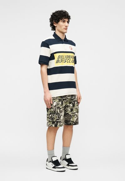 Νεαρός άνδρας που φοράει μπλούζα πόλο σε ναυτικό μπλε και λευκές ρίγες με το κείμενο "Billionaire Boys Club", καμουφλάζ cargo σορτς, γκρι κάλτσες και ασπρόμαυρα αθλητικά παπούτσια.
