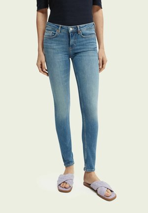 Femme portant un jean slim bleu clair et des sandales matelassées lavande, debout devant un fond uni.