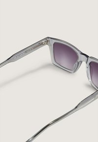 Duidelijke plastic zonnebril met een gedurfde, hoekige rand en verloopgrijze lenzen. Accenten van zilverkleurig metaal op de slapen.