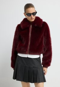 Burgundská umělá kožešinová bomber bunda se zipem vpředu, manžetami a límcem s žebrováním. Kombinována s bílou blůzkou a černou skládanou sukní.
