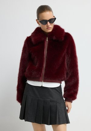 Veste bomber en fausse fourrure bordeaux avec fermeture éclair à l'avant, poignets et col côtelés. Associée à un chemisier blanc et une jupe plissée noire.