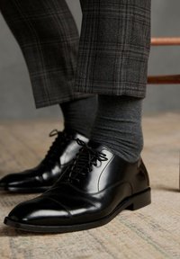 Los zapatos de vestir de cuero negro cuentan con un acabado suave, punta redonda y cordones finos. Combinados con calcetines gris oscuro y pantalones de cuadros.