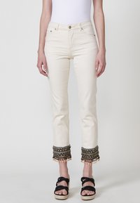 Jeans de denim color crema con acentuaciones de cuentas y flecos en los puños, que presentan un diseño clásico de cinco bolsillos y cierre con cremallera.